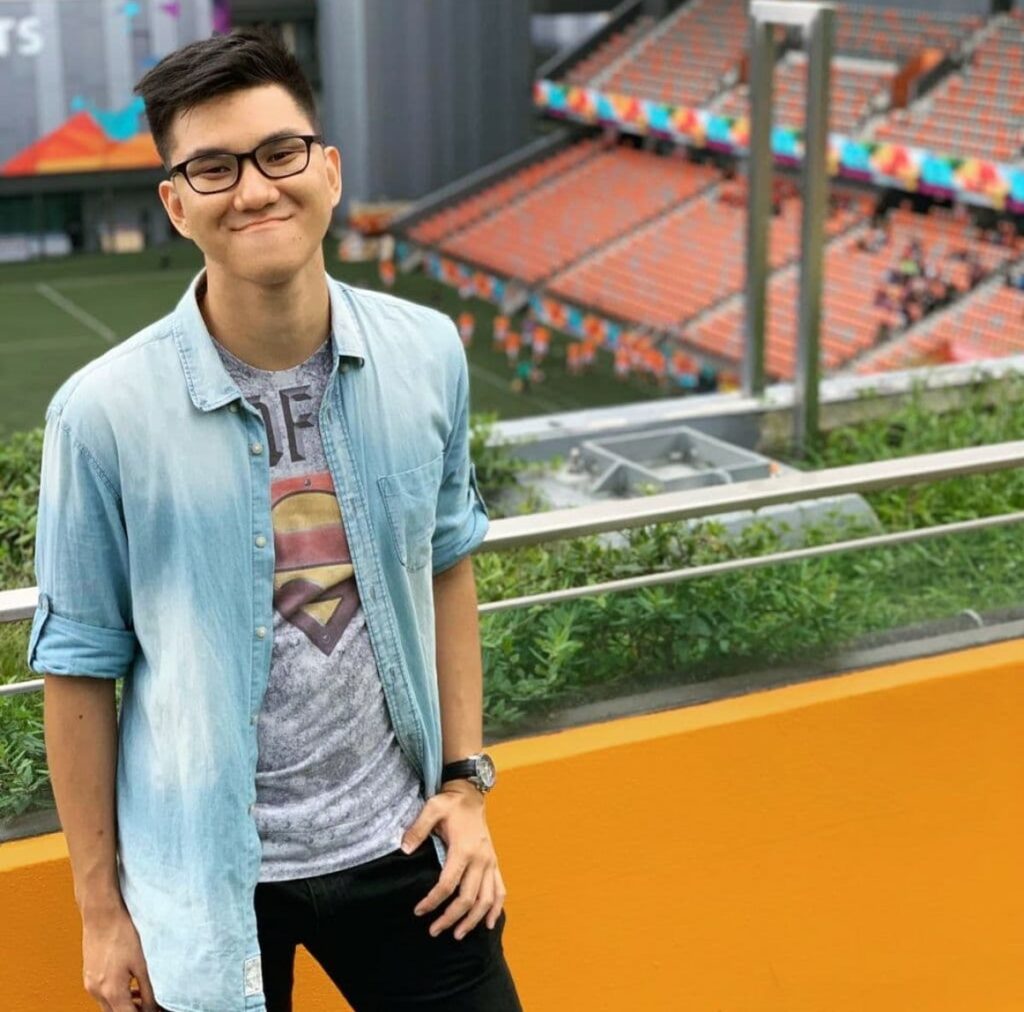 Bjorn in a blue shirt worn over a grey t-shirt, standing against a backdrop of a stadium