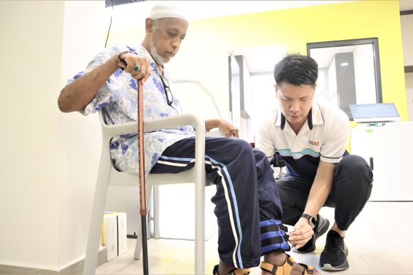 Power of Care – Holistic Care Through Collaboration 2 A senior holding a walking stick seated in a chair while Winson kneels beside him to fasten a leg weight strap around his lower leg.