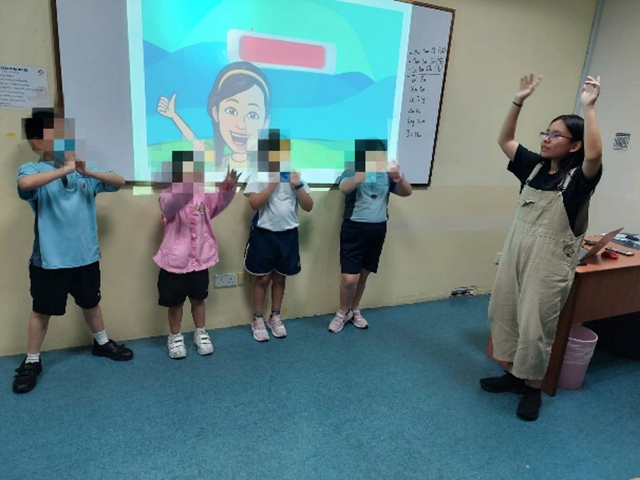 Cindy as a volunteer teaching assistant leading a group of 4 children in school uniforms in a song session
