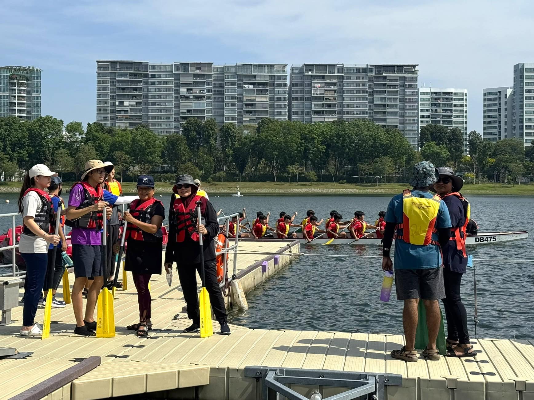 Strength in Community: She Came to Help Others and Found Support Herself 1 Sindhu with other volunteers and E S H clients, all in life jackets, waiting at the dock. A dragonboat propelled by another team races across in the background.