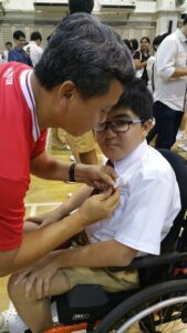 Mr Sim helping Aaron to pin his name tag on his school uniform in a school hall